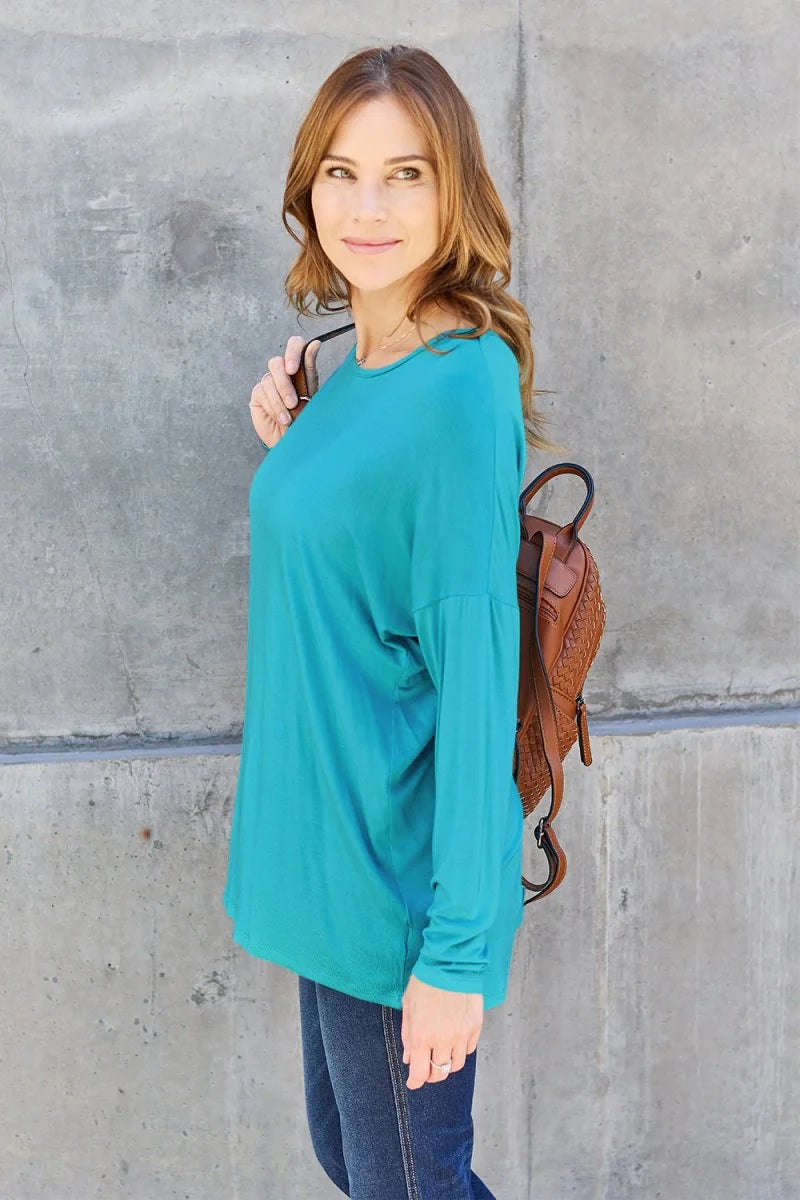 A woman wearing a teal long-sleeve top and dark wash jeans, with a brown woven backpack.