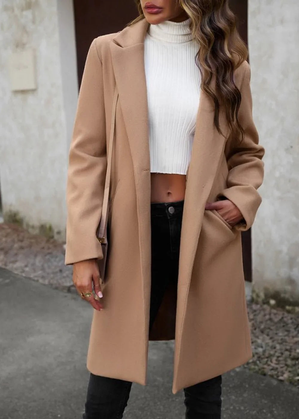 Woman in a tan wool-blend coat over a white ribbed turtleneck crop top, black jeans, and gold rings.