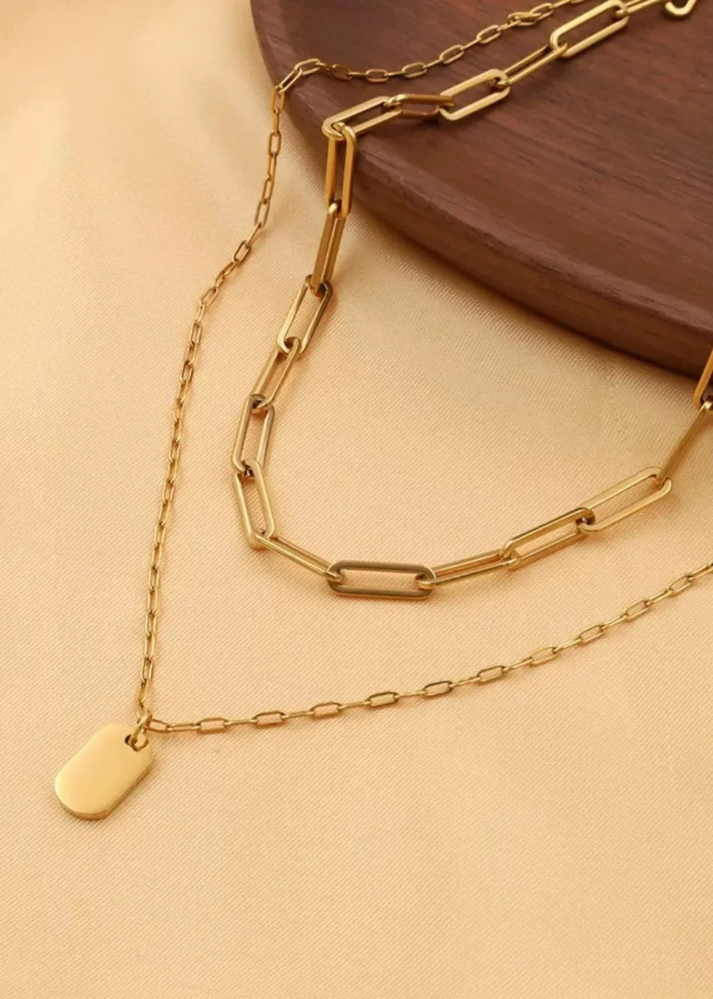 Gold-plated layered necklace with a small oval tag pendant on a light peach textured background.
