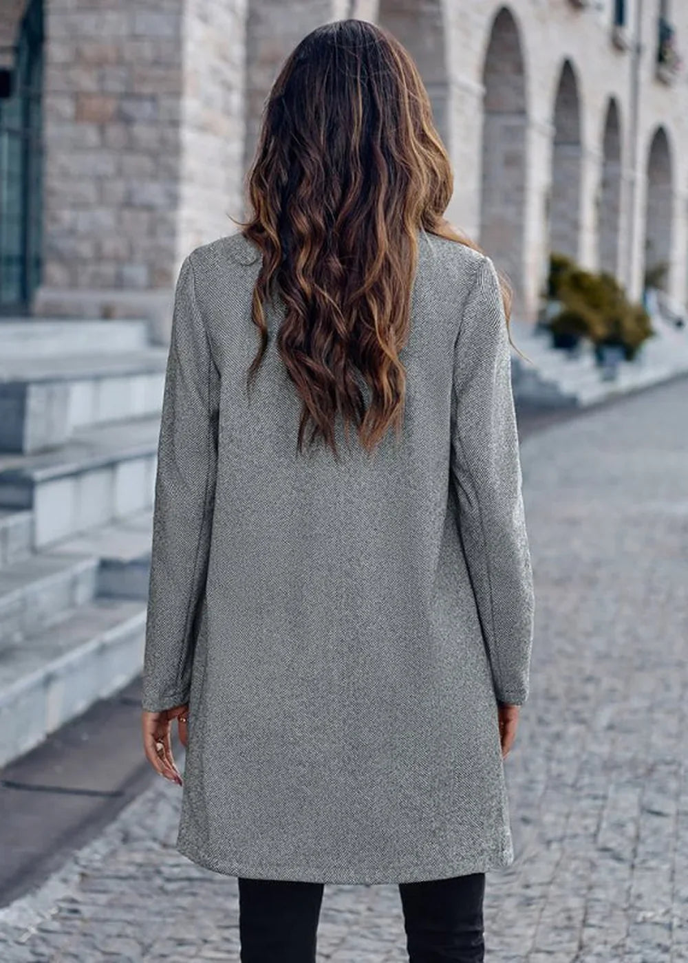 Back view of a woman wearing a textured grey jacket with long sleeves and black pants.