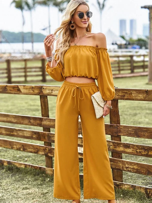 Woman in mustard yellow off-the-shoulder crop top and wide-leg pants, holding a woven clutch.