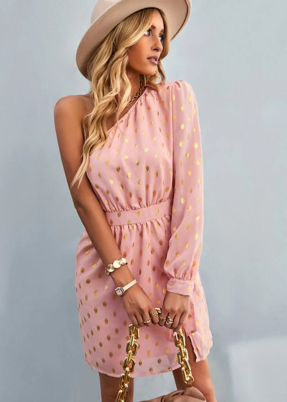 Pink one-shoulder mini dress with gold polka dots, cinched waist, and gold chain bag.