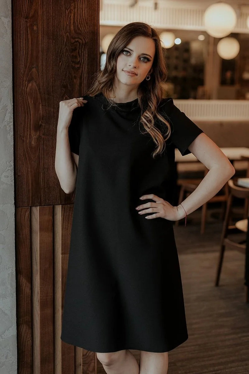 Loose-fit black short-sleeve day dress with a subtle sheen, worn by a model against a wood-paneled background.