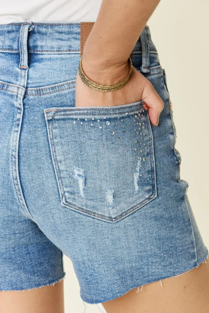 Light wash distressed denim shorts with rhinestone pocket detail and frayed hem.