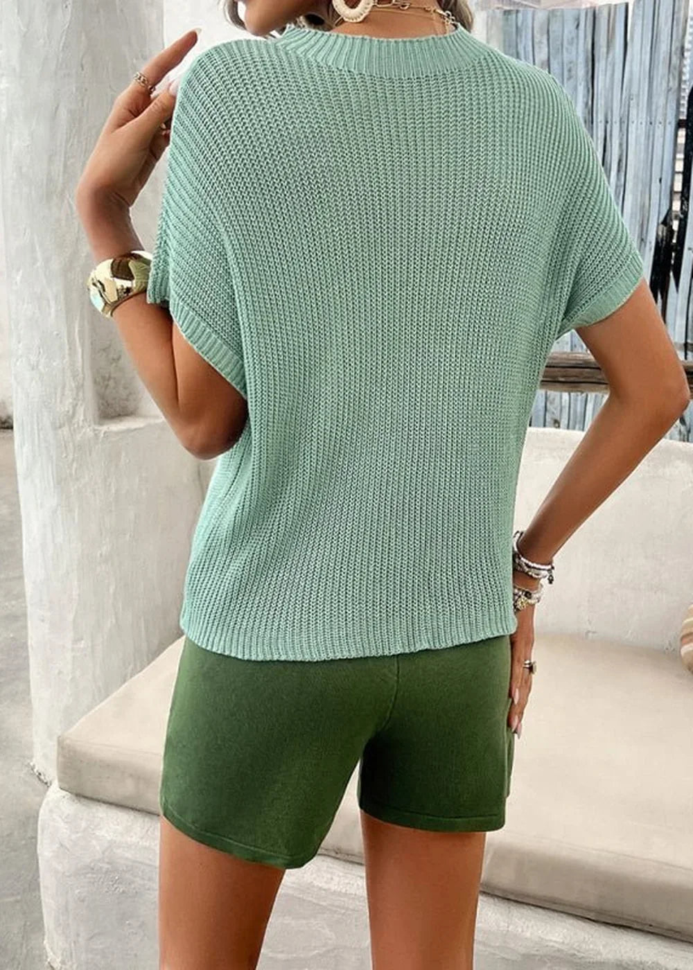Back view of a woman wearing a light green knit short-sleeve top and dark green shorts.