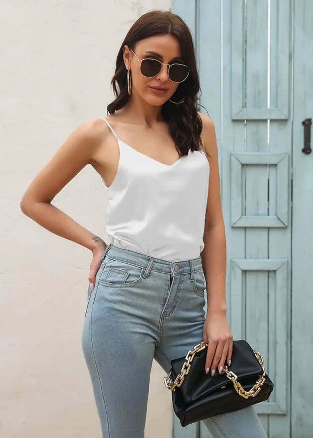 Woman in white satin cami and light wash jeans holding a black clutch with gold chain strap.