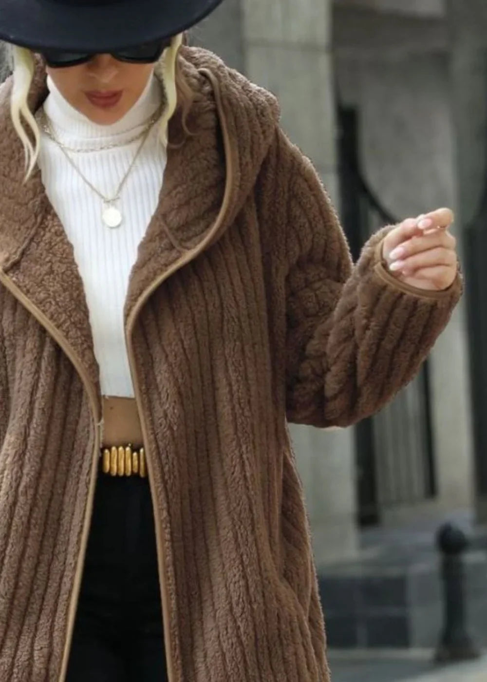 Plush brown hooded coat with ribbed texture and tan trim, worn over a white turtleneck and black pants.