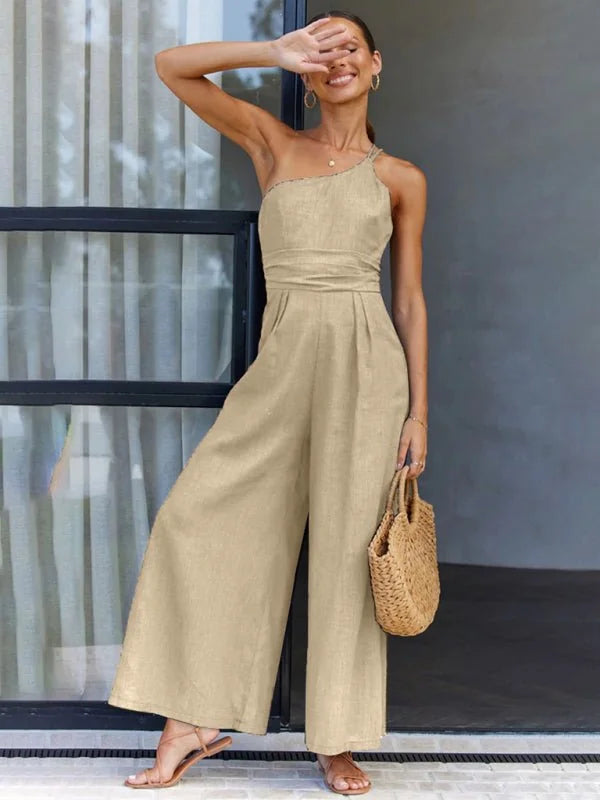 Beige one-shoulder wide-leg jumpsuit with tie waist and straw bag, on model.