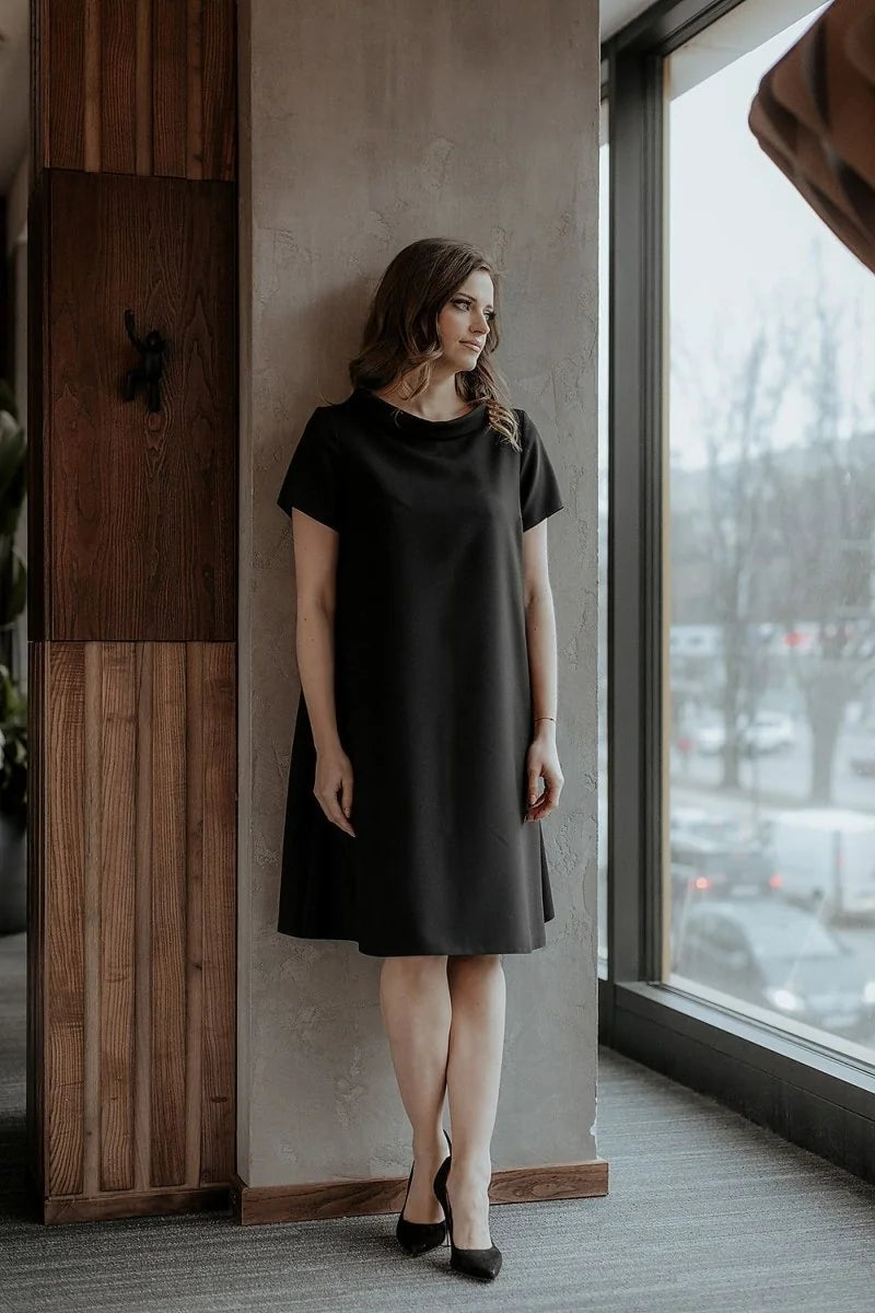 Woman in a loose-fit black dress with short sleeves, standing by a window.