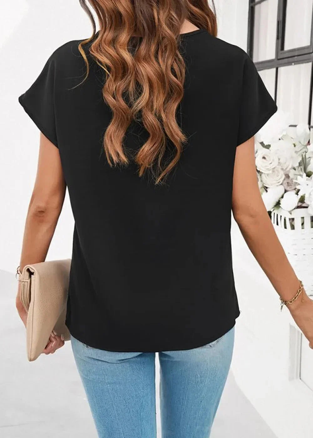 Back view of a woman wearing a black short-sleeve top and light blue jeans, holding a beige clutch.