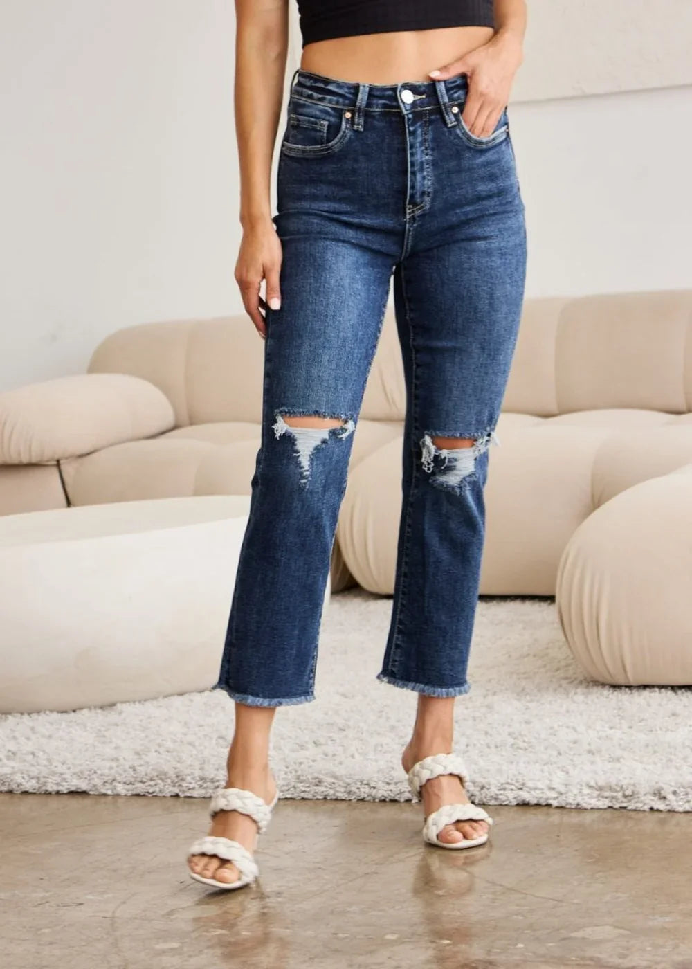 Dark wash distressed denim jeans with frayed hem and white braided sandals on model.