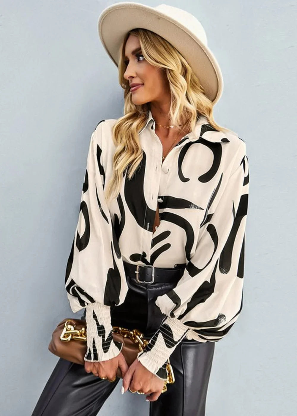 Woman wearing a white and black abstract print button-down shirt with voluminous sleeves, black pants, and a cream hat.