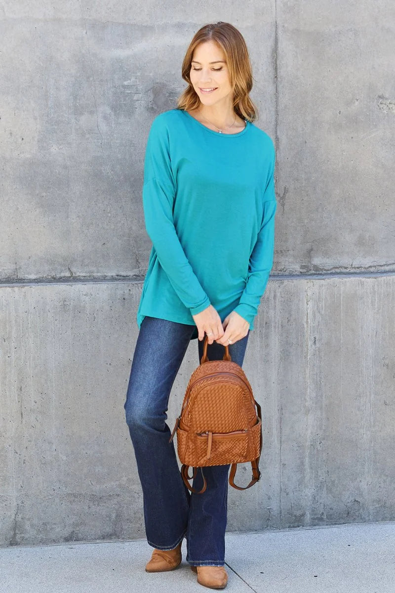Teal long-sleeve top with relaxed fit, paired with dark wash flare jeans and a woven brown backpack.
