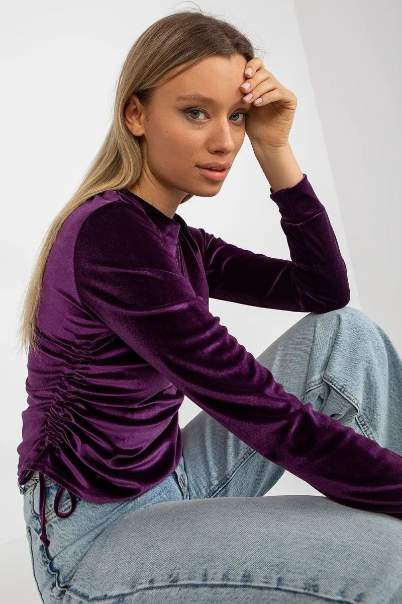 Purple velvet long-sleeve top with ruched side detail, worn with light wash denim jeans.