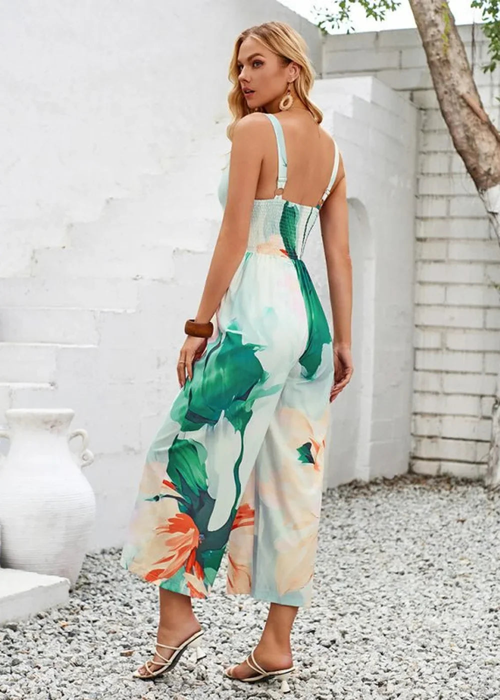 Woman in a flowing, light blue jumpsuit with a large abstract floral print in green and orange, featuring a smocked bodice...