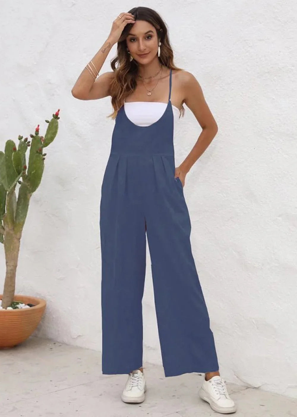 A woman models a navy blue wide-leg jumpsuit with a white tank top underneath, paired with white sneakers.