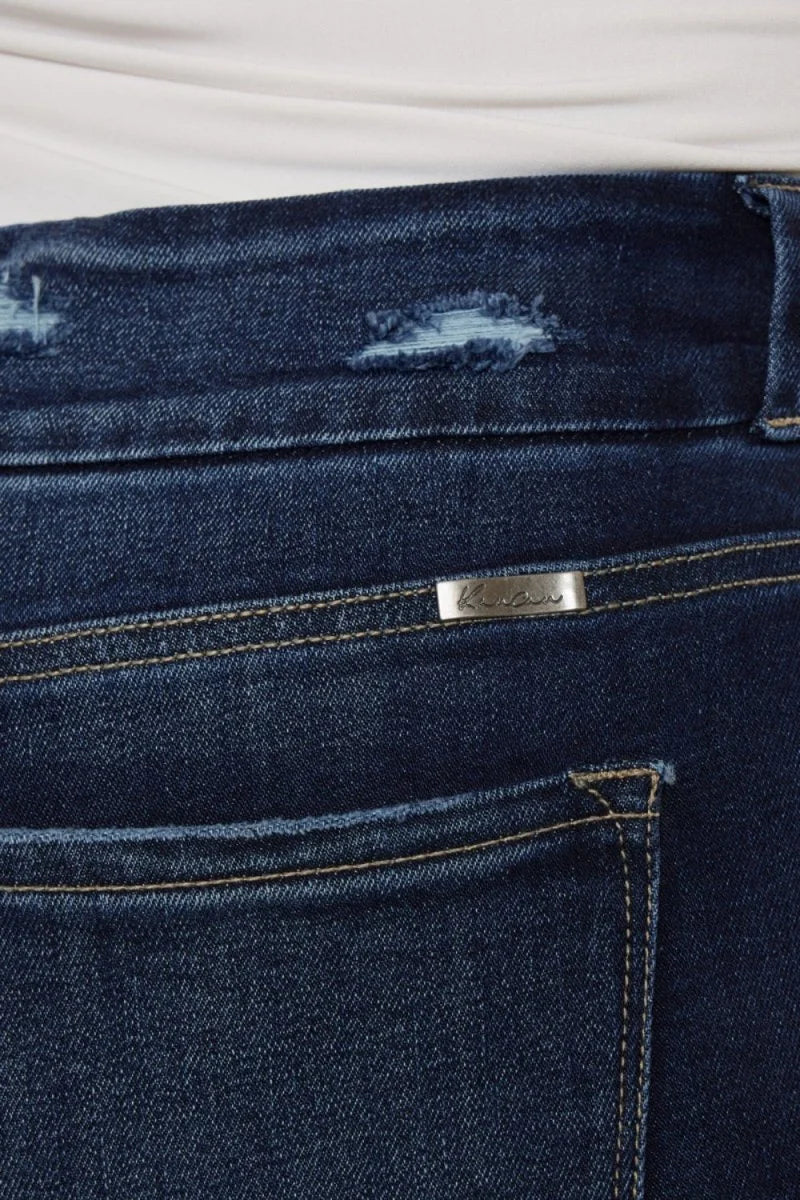 Close-up of dark wash distressed denim with a silver logo plate and frayed hem detail.