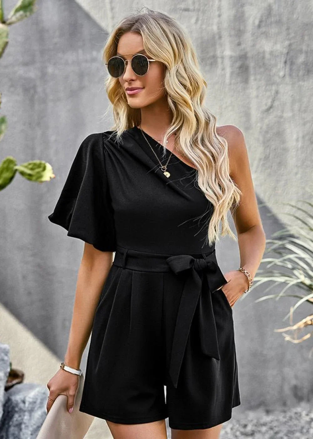 Black one-shoulder romper with flutter sleeve and tie waist, on model.
