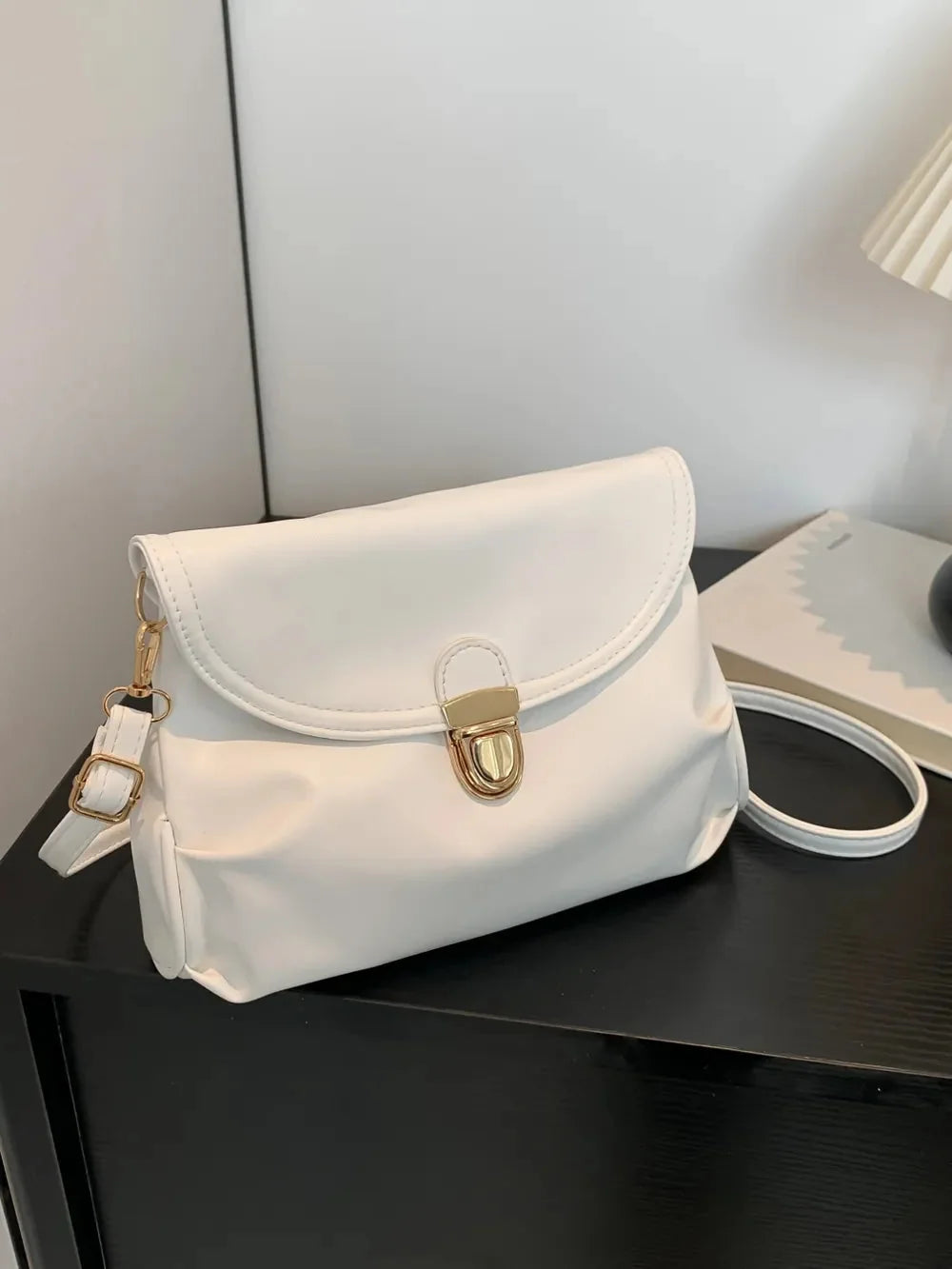 White faux leather crossbody bag with gold hardware and adjustable strap, on a black surface.