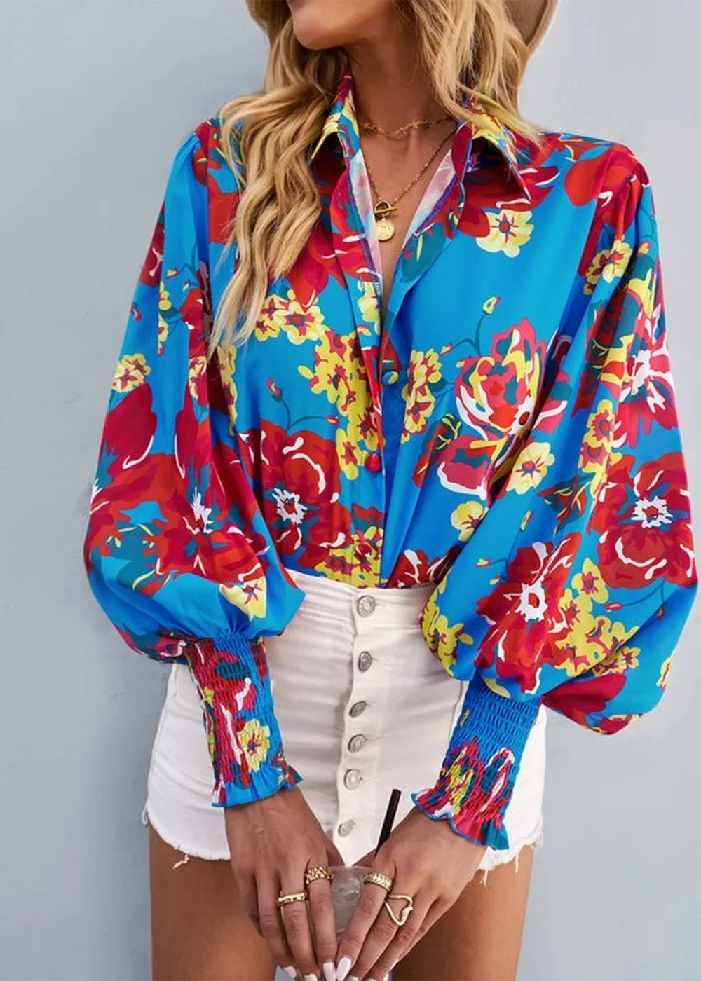 Blue long-sleeve blouse with a vibrant floral print and cinched cuffs, worn by a model with white shorts.