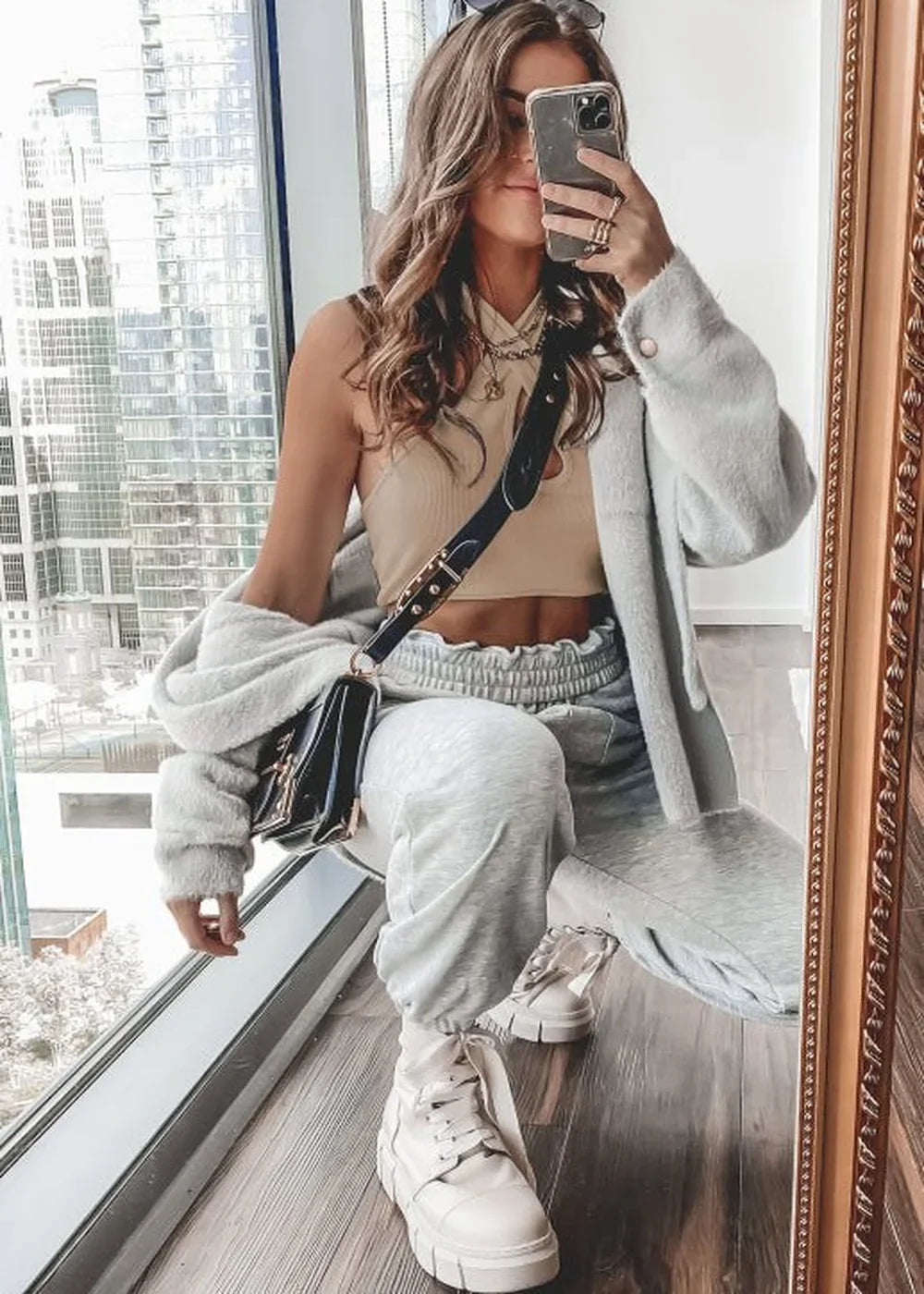 Woman in a cream ribbed tank top and light gray sweatpants, holding a black purse and taking a mirror selfie.