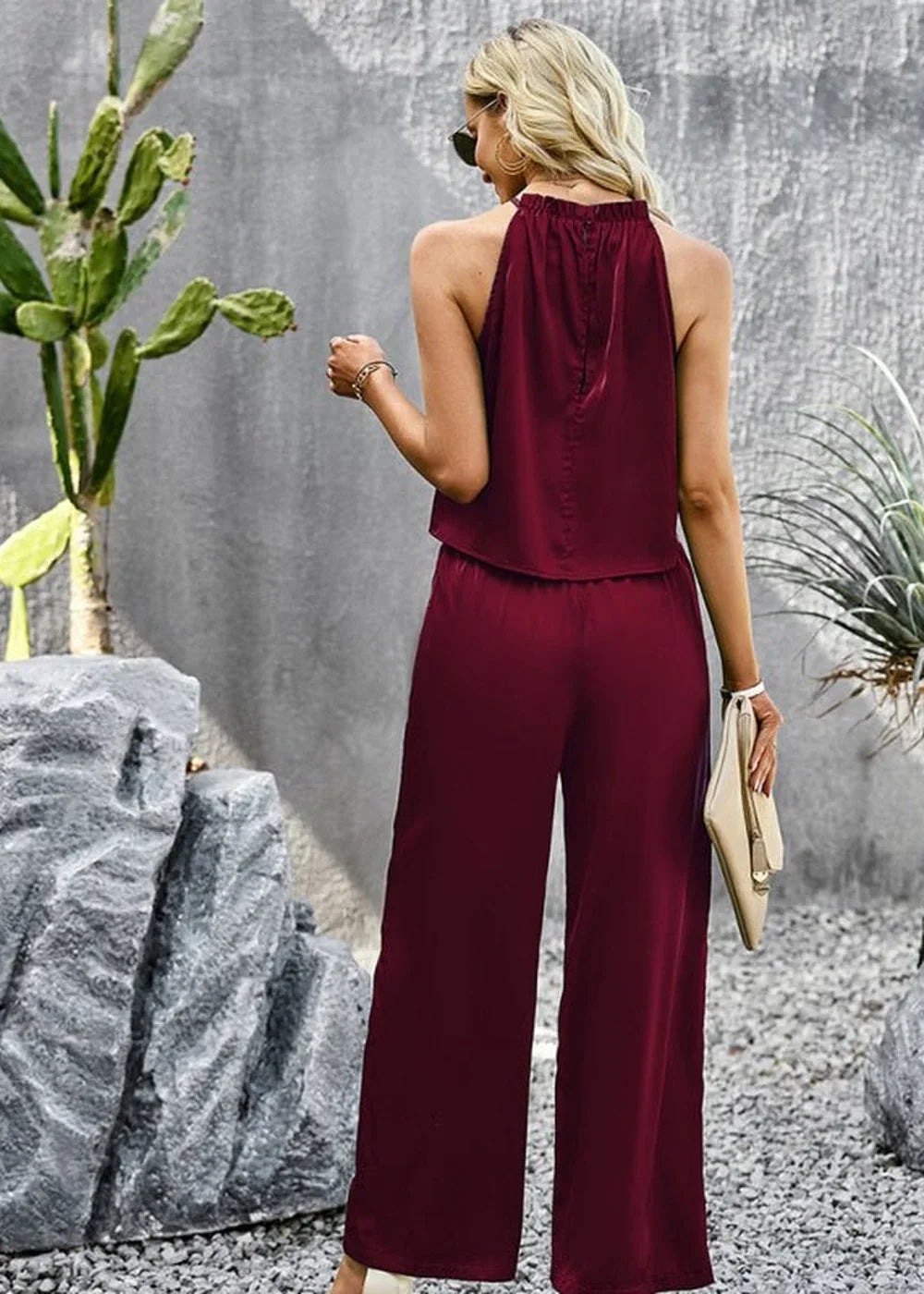Woman in a maroon sleeveless jumpsuit with wide legs, holding a cream clutch.