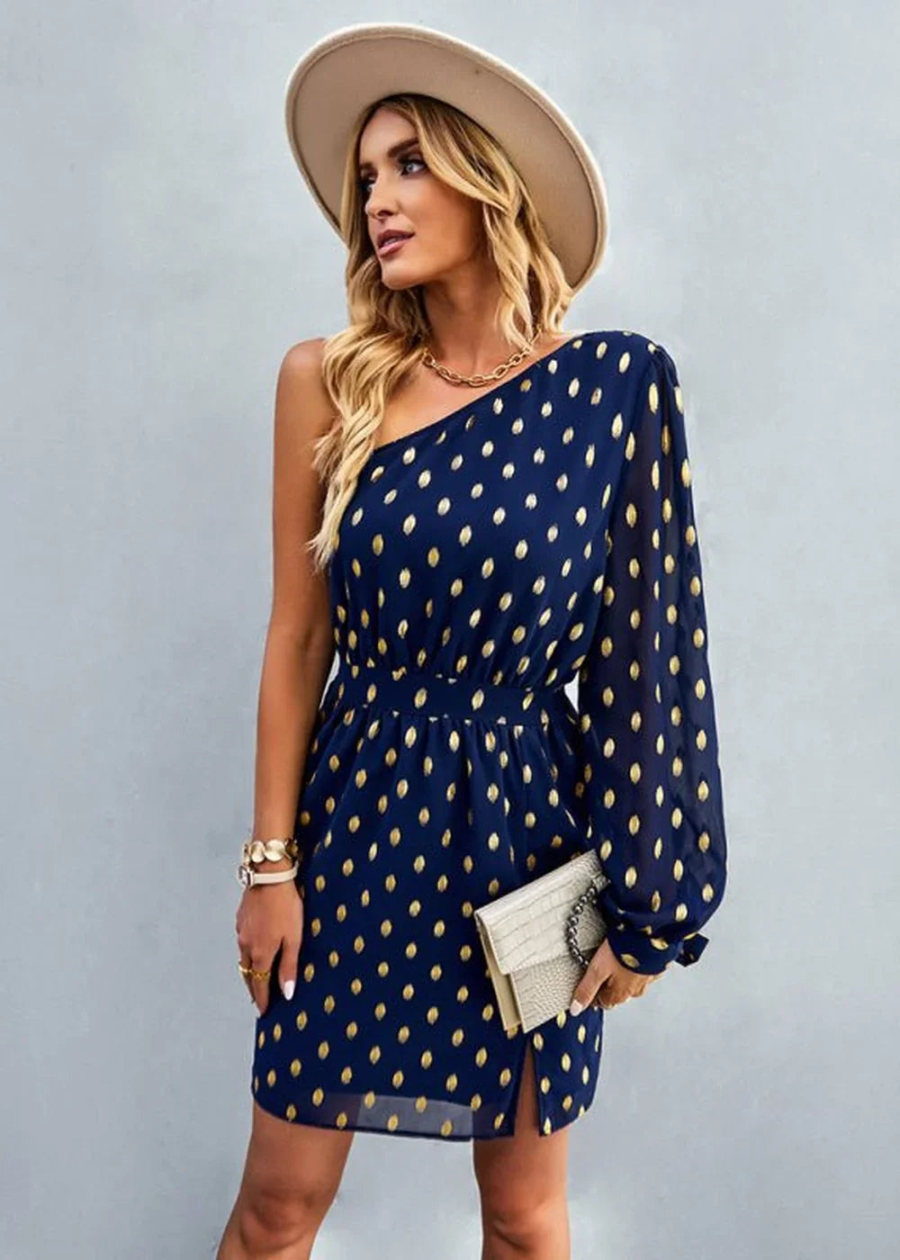 Navy blue one-shoulder dress with gold polka dots, cinched waist, and a light-colored clutch.