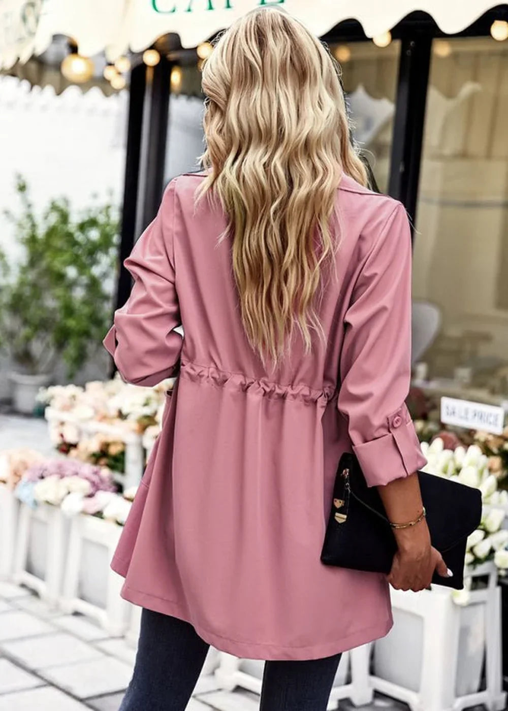 Back view of a woman wearing a pink trench coat with a cinched waist and rolled sleeves, holding a black clutch.
