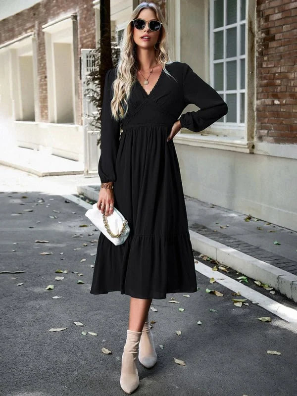 Black tiered midi dress with V-neck and long sleeves, held by a woman wearing neutral booties and a white clutch.
