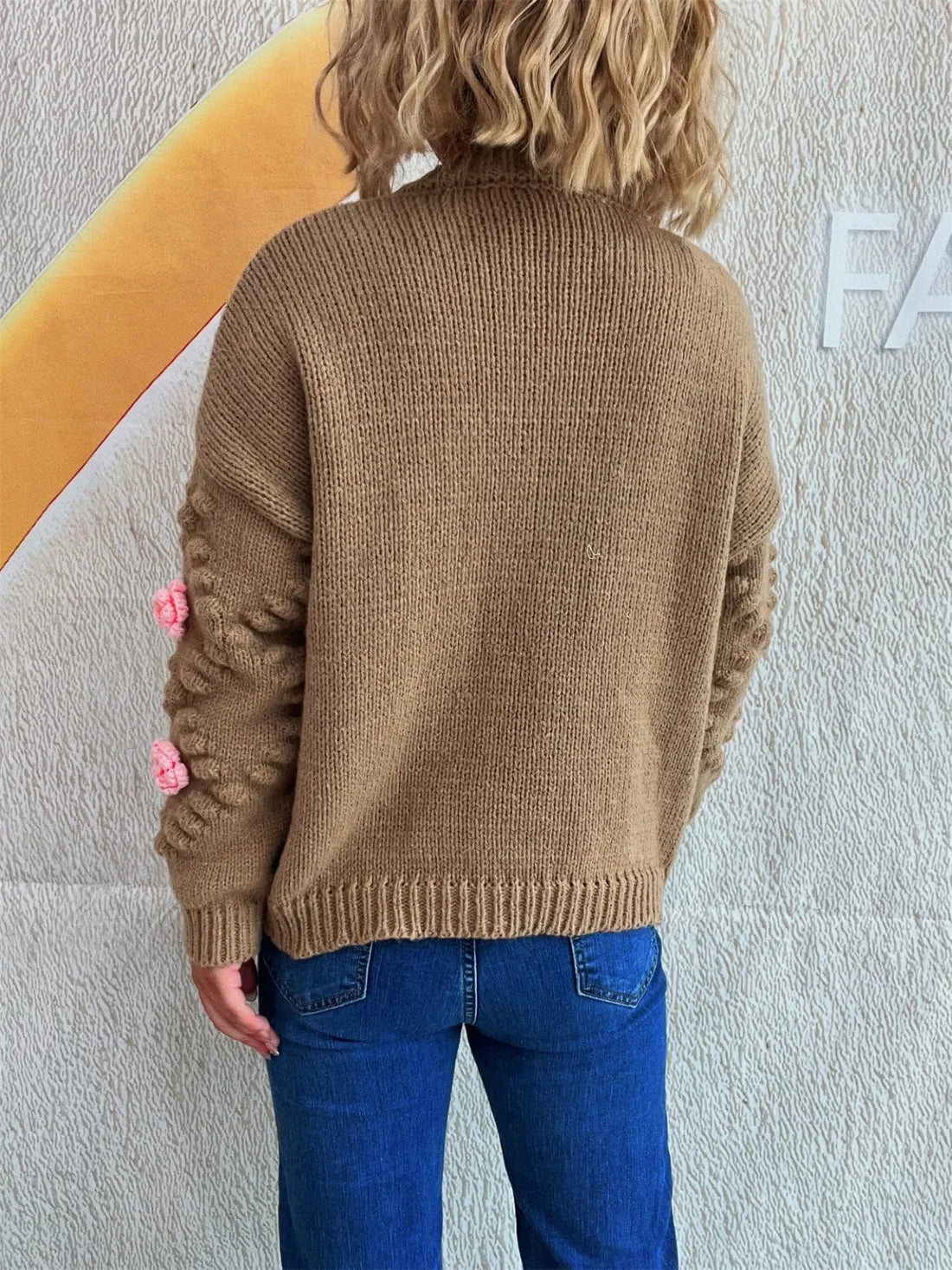 Brown knit sweater with textured sleeves and pink floral accents, worn by a model with blue jeans.