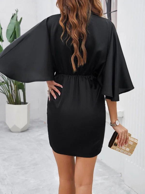 Black satin mini dress with dramatic flutter sleeves, cinched waist, and a gold clutch.