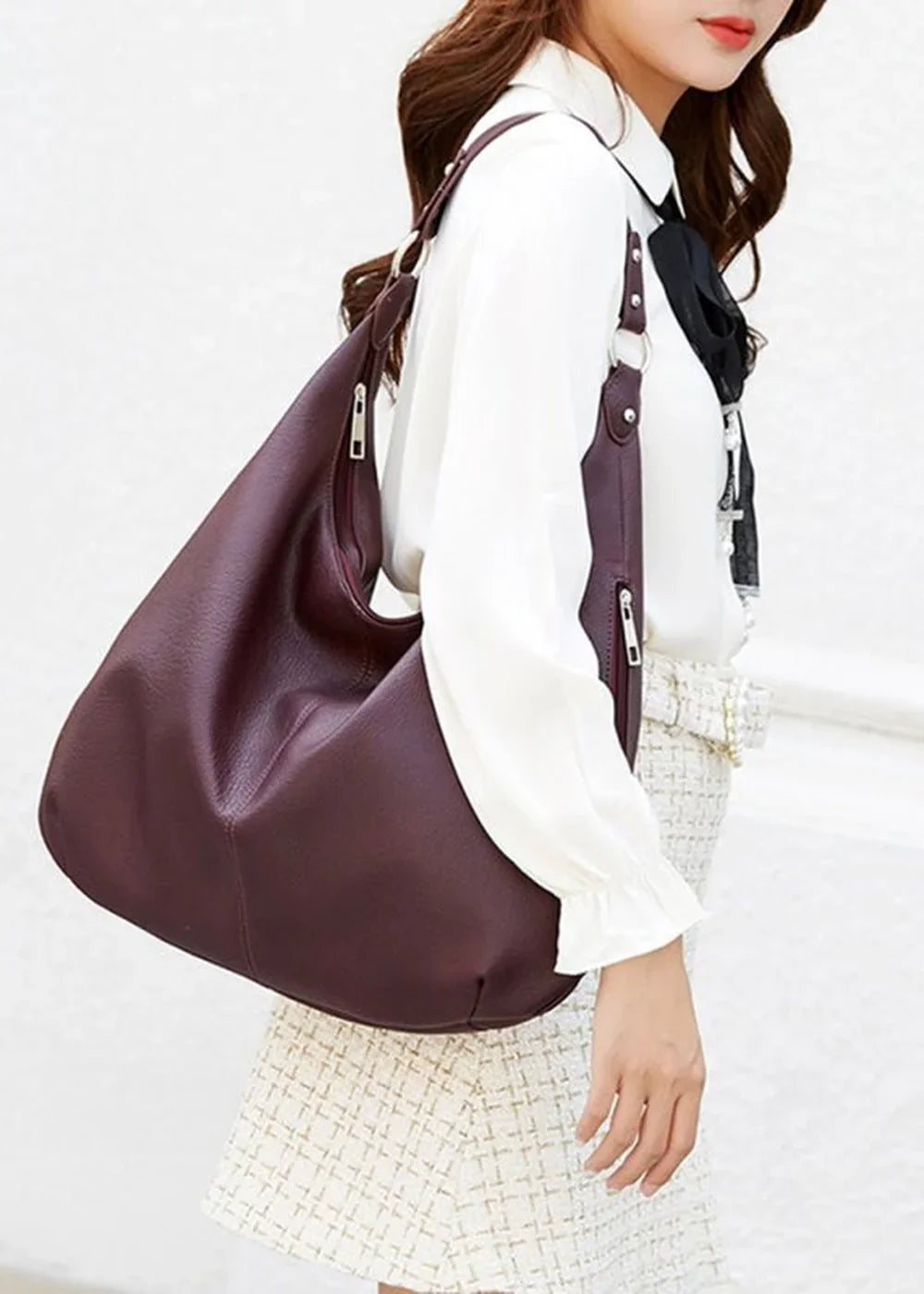Deep burgundy faux leather hobo bag with silver hardware, worn by a woman in a white blouse and textured skirt.