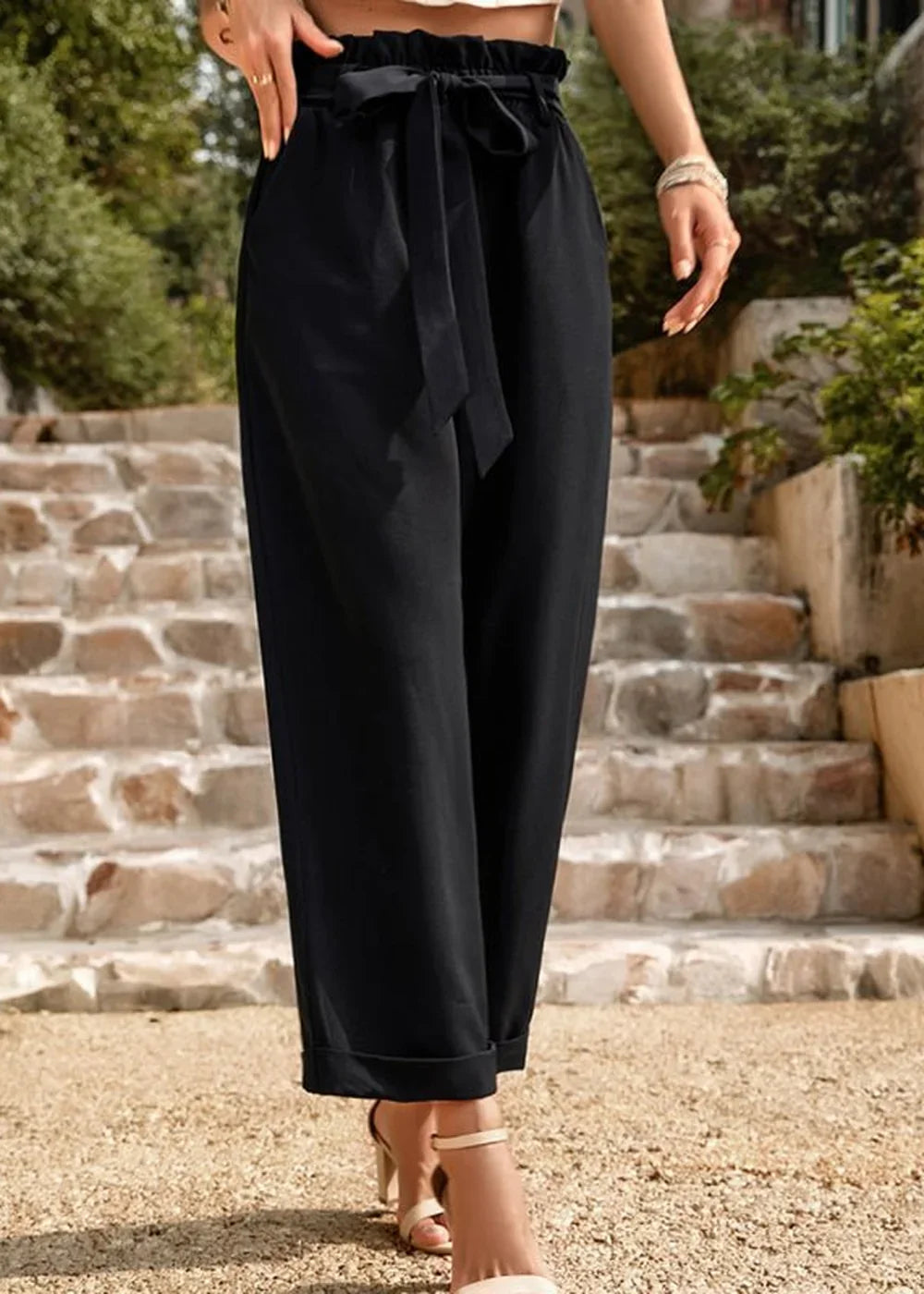 Black paperbag waist pants with a self-tie belt, shown on a model standing on stone steps.