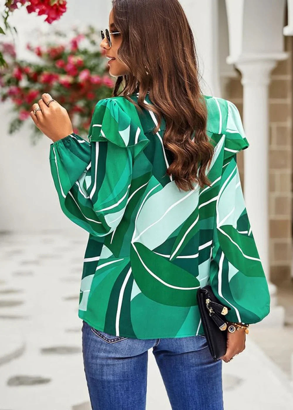 Woman in a green floral print blouse with ruffled shoulders and blue jeans, holding a black clutch.