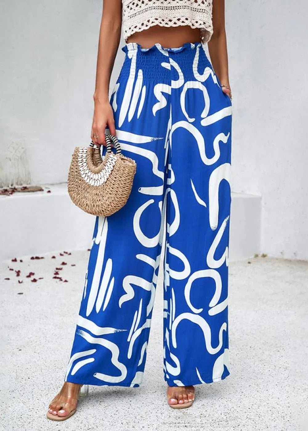 Blue wide-leg pants with a white abstract print and a smocked waistband, worn by a model holding a straw bag.