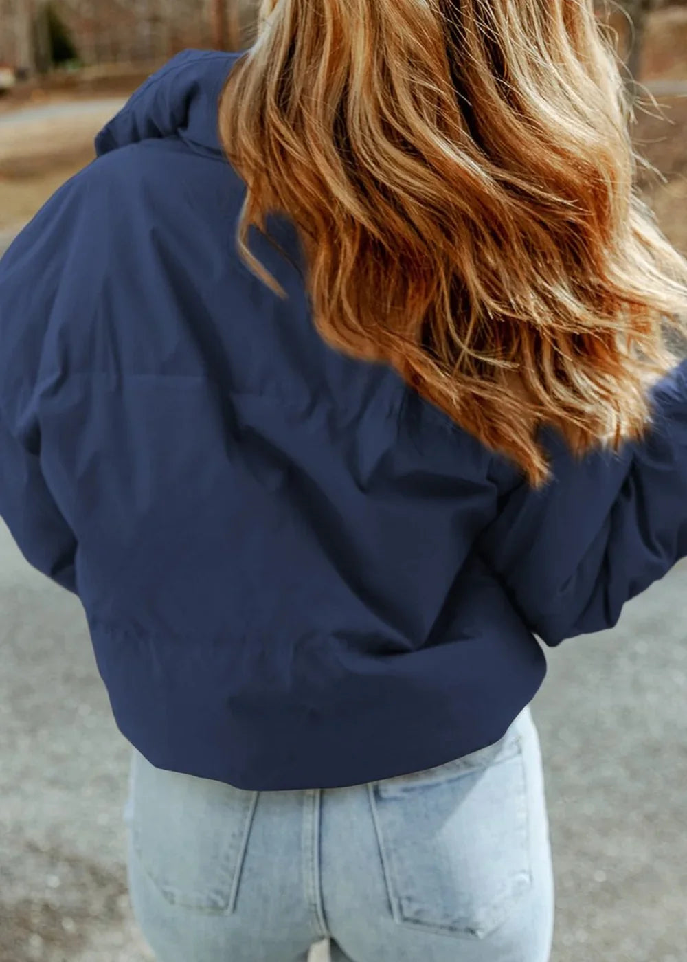 Back view of a navy blue puffer jacket on a woman with long, wavy blonde hair, paired with light wash jeans.