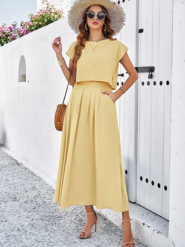 Woman in a pale yellow two-piece set with a cropped top and pleated midi skirt, wearing a straw hat and nude heels.