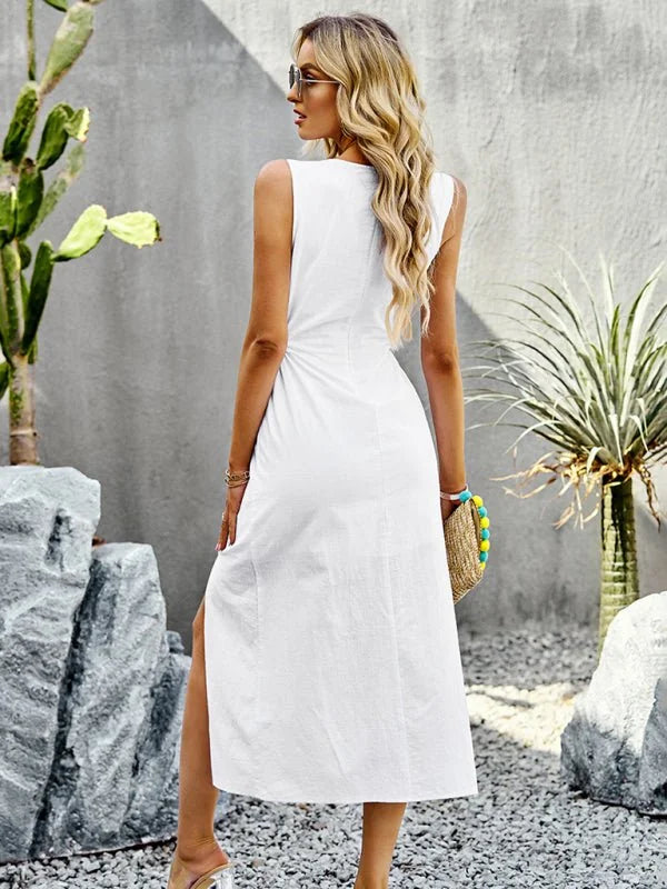 Woman from behind in a white sleeveless midi dress with a high slit, holding a straw bag with colorful pom-poms.