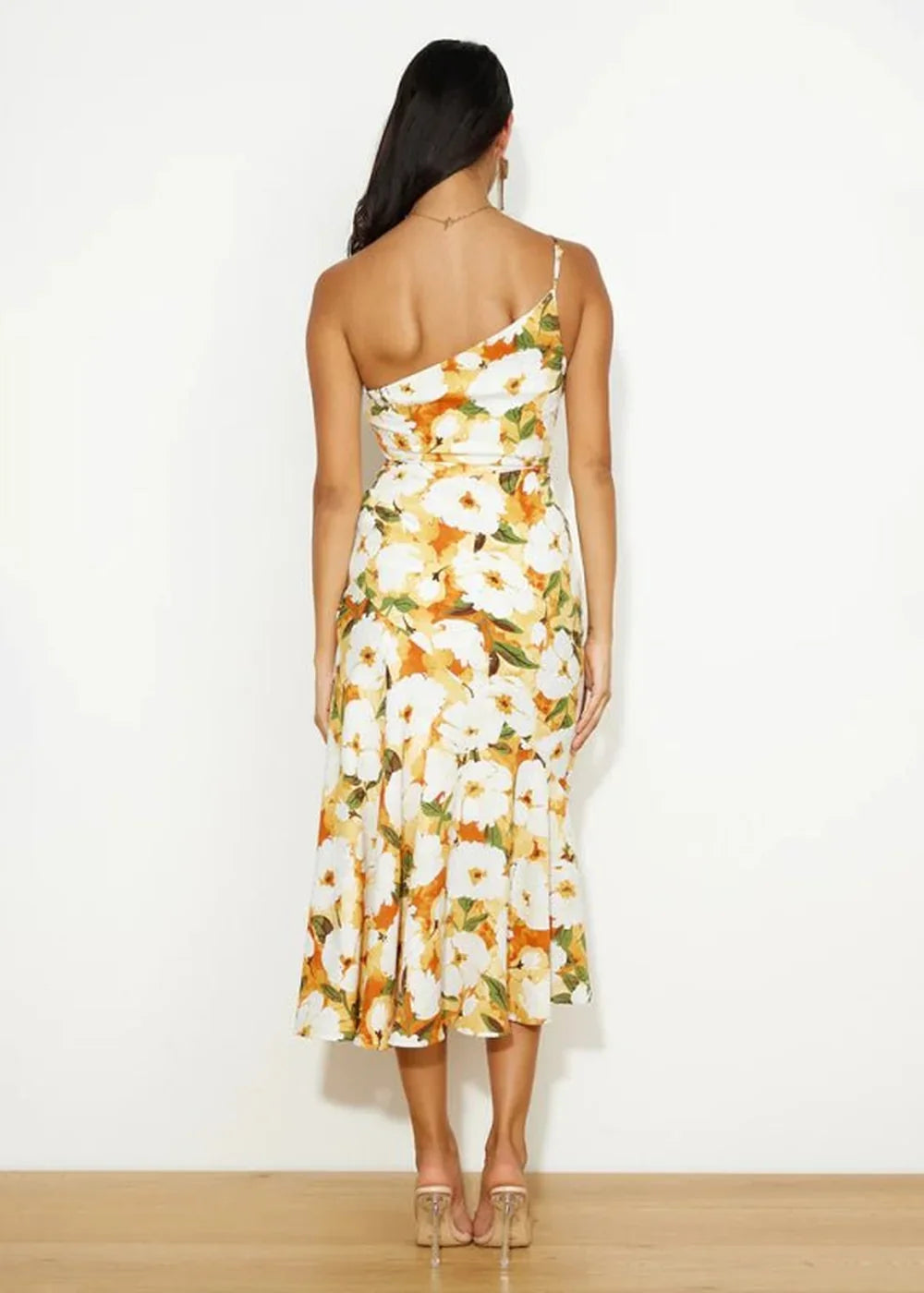 Back view of a woman in a white one-shoulder midi dress with a bold yellow, orange, and green floral print.