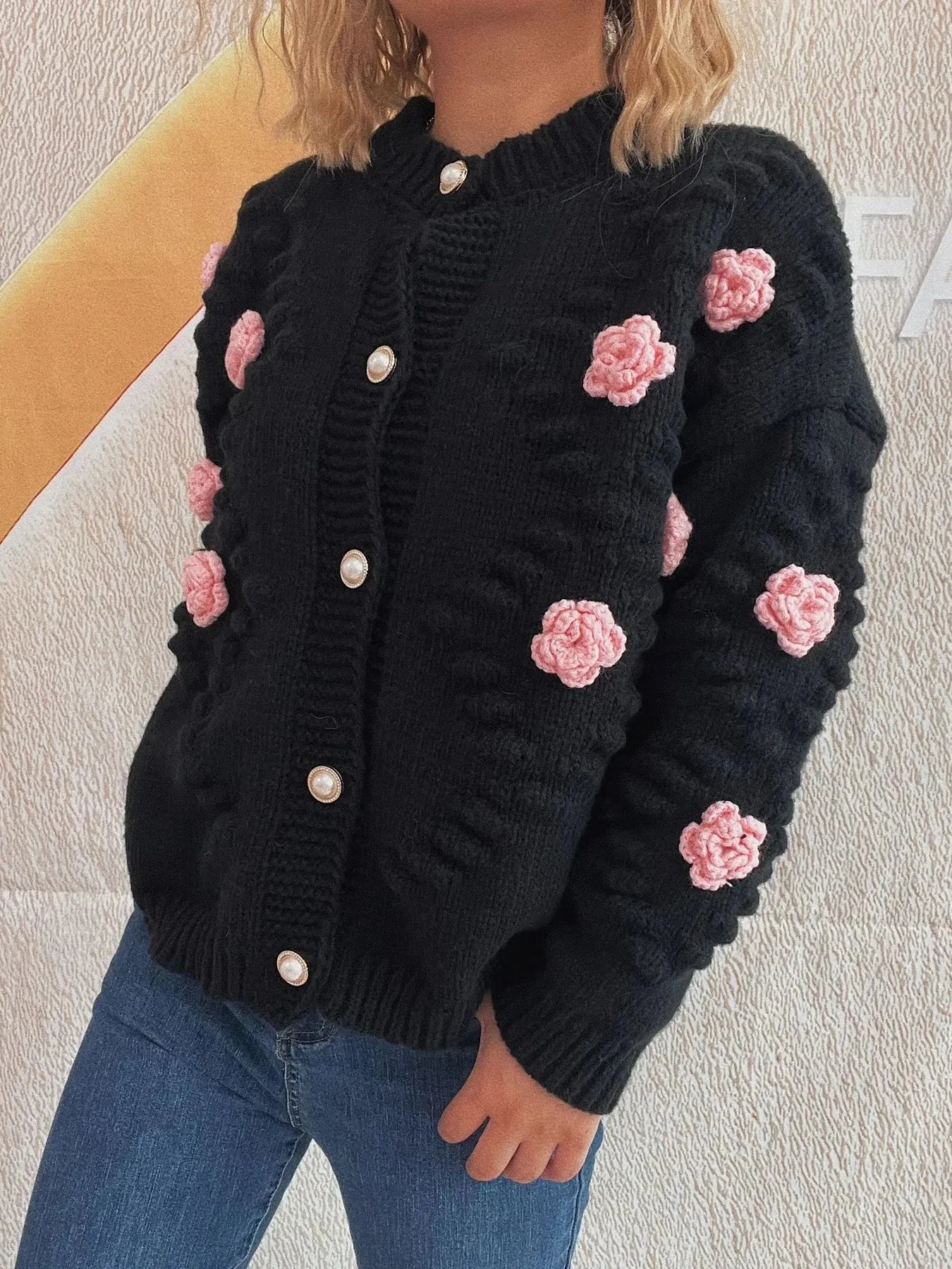 Black knit cardigan with pink floral appliques and pearl buttons, worn over blue jeans.
