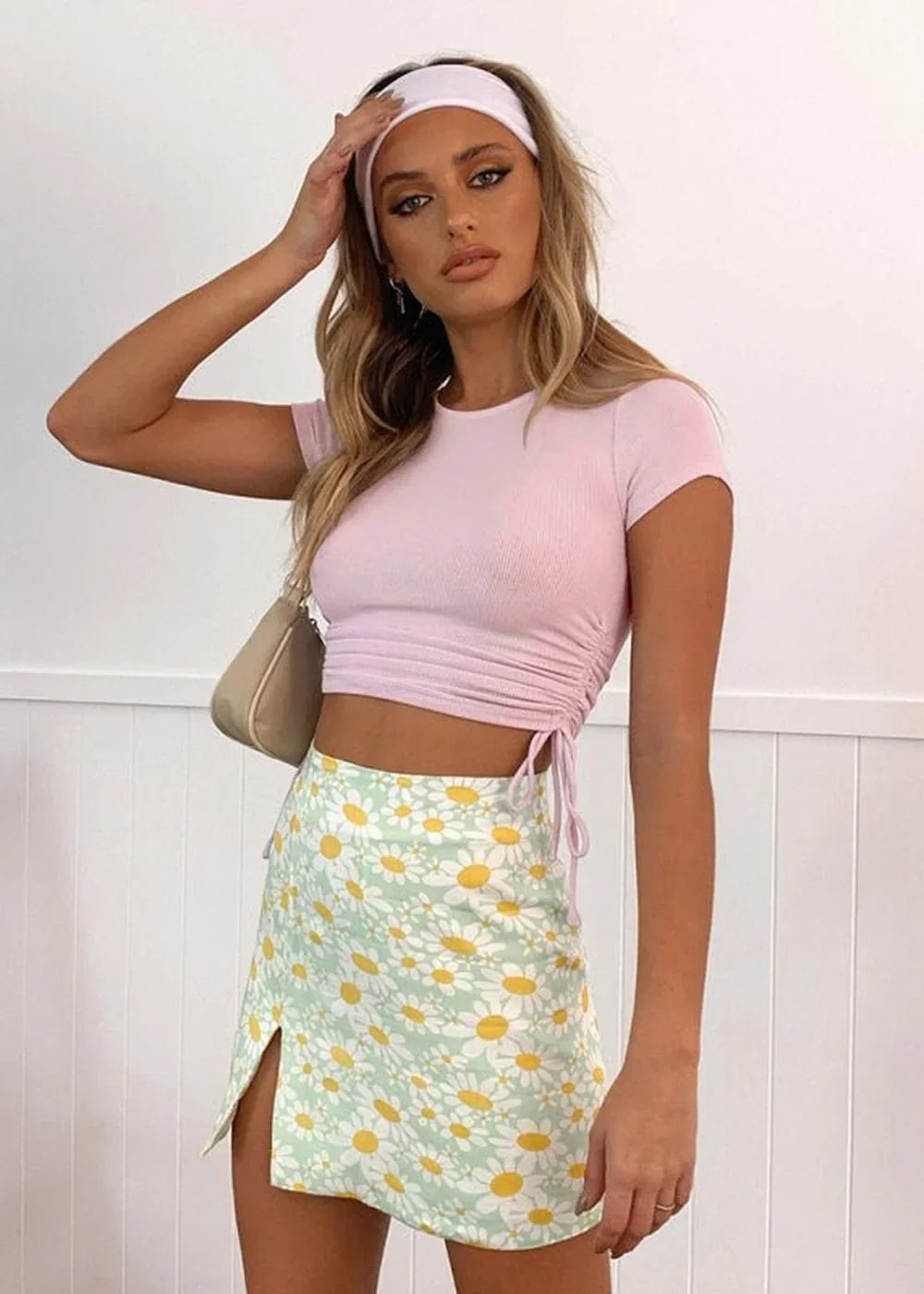 A woman in a pink ruched crop top and a light green daisy-print mini skirt with a thigh slit.