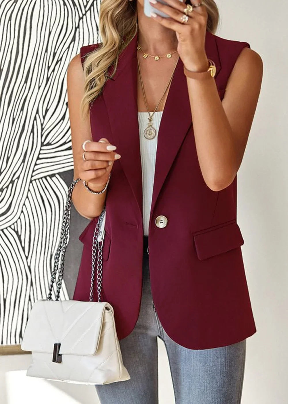 Burgundy sleeveless blazer with a white quilted handbag and gold jewelry.