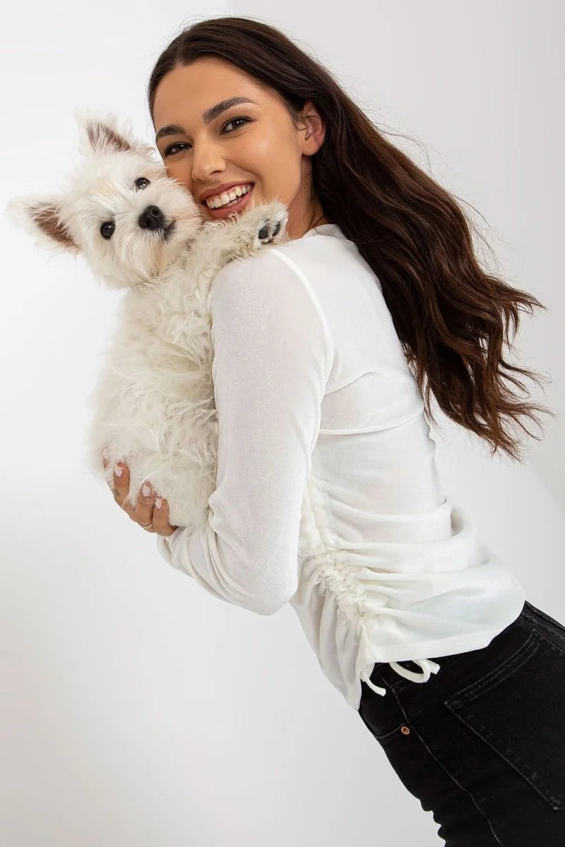 Woman in a white ruched long-sleeve top and black jeans holding a fluffy white dog.