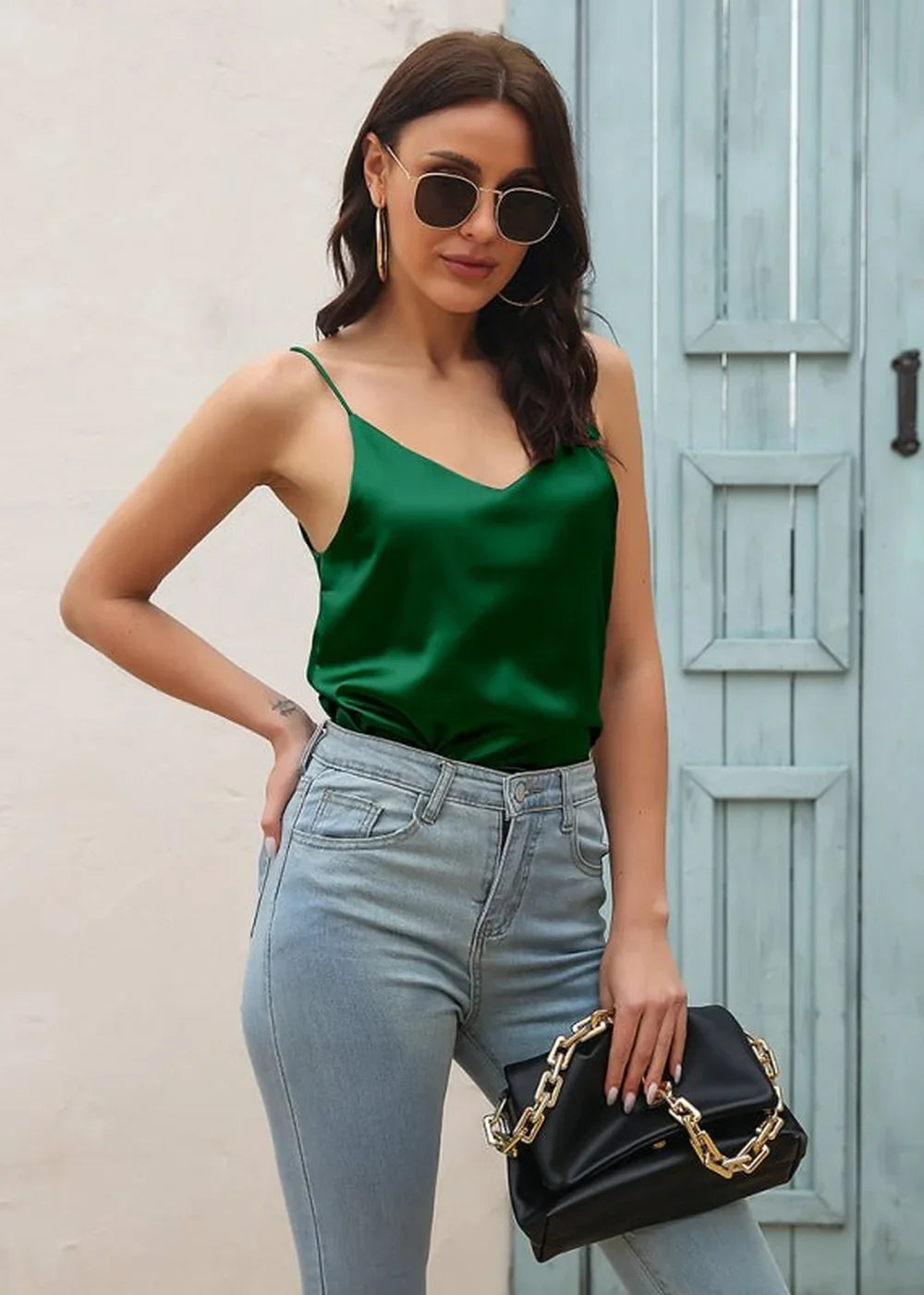Woman in a shiny emerald green V-neck camisole and light wash jeans, holding a black clutch with a gold chain.