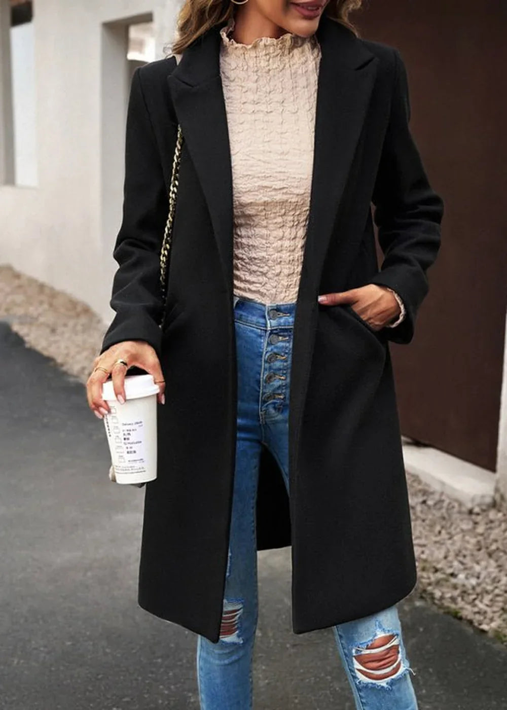 Black wool-blend coat with a notched lapel and gold chain detail, worn over a textured tan top and distressed jeans.