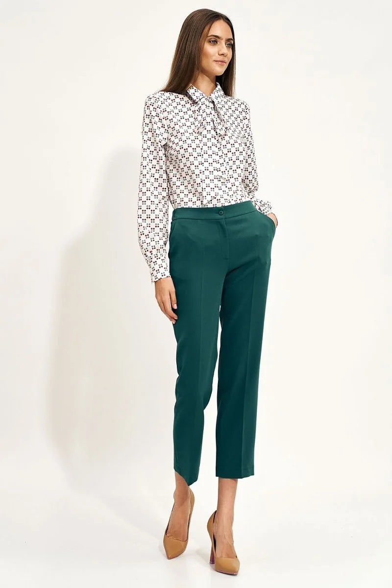 Full-length shot of a woman wearing a white patterned blouse and dark teal, straight-leg, cropped pants.