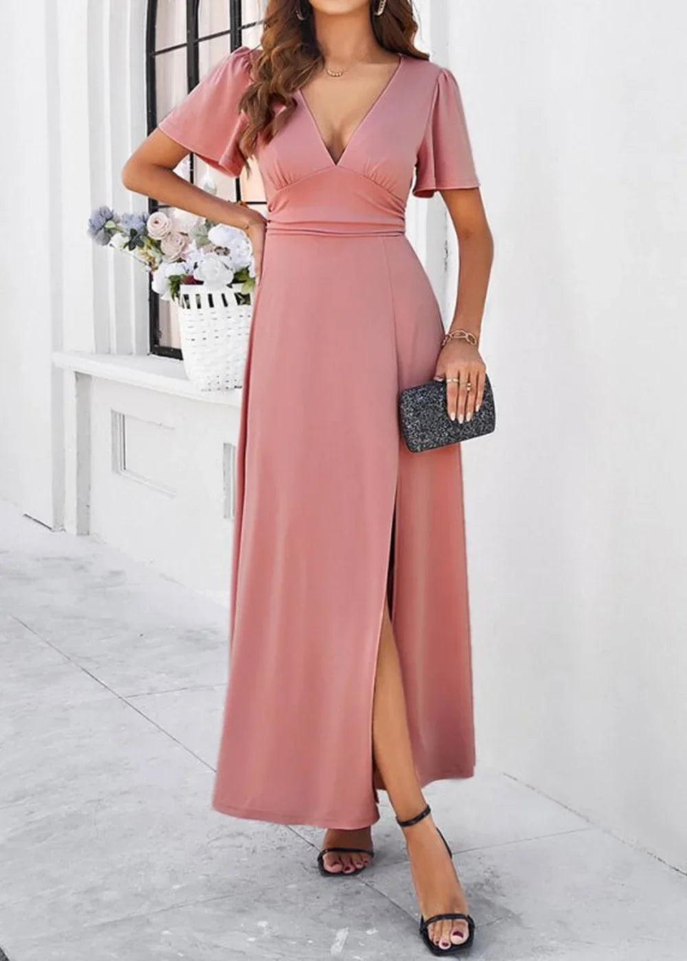 Pink maxi dress with flutter sleeves, V-neck, and thigh-high slit, worn with black heels and a sparkly clutch.
