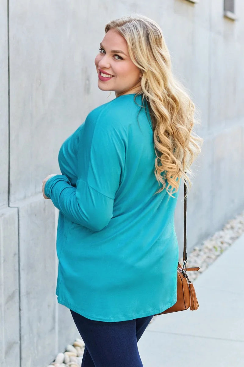 Turquoise long-sleeved top with a relaxed fit, worn by a model with blonde wavy hair, carrying a brown crossbody bag.