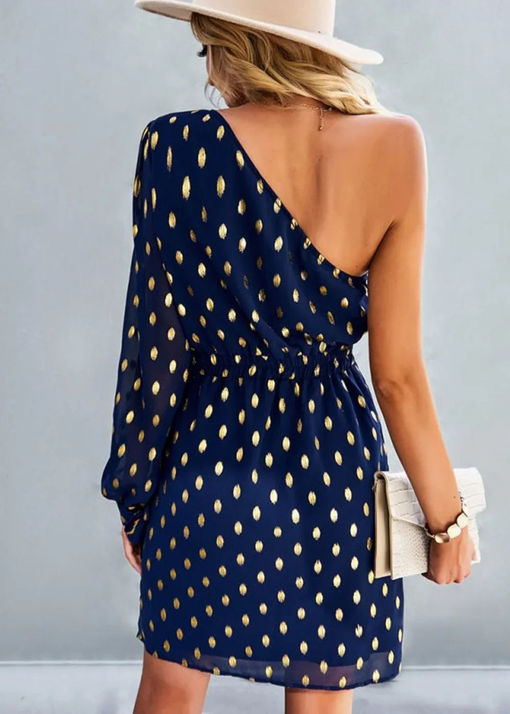 Navy one-shoulder dress with gold polka dots, cinched waist, and long sleeve.