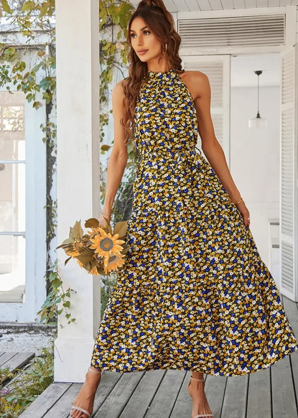 Woman in a black halter maxi dress with a yellow, white, and blue floral print, holding sunflowers.